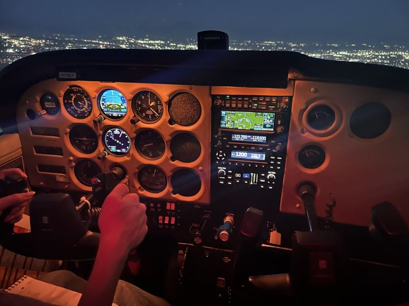Night cockpit view of a lit instrument panel with a Garmin GPS and radio stack, an instructor's and a student's hands on the yoke, and city lights outside the windshield